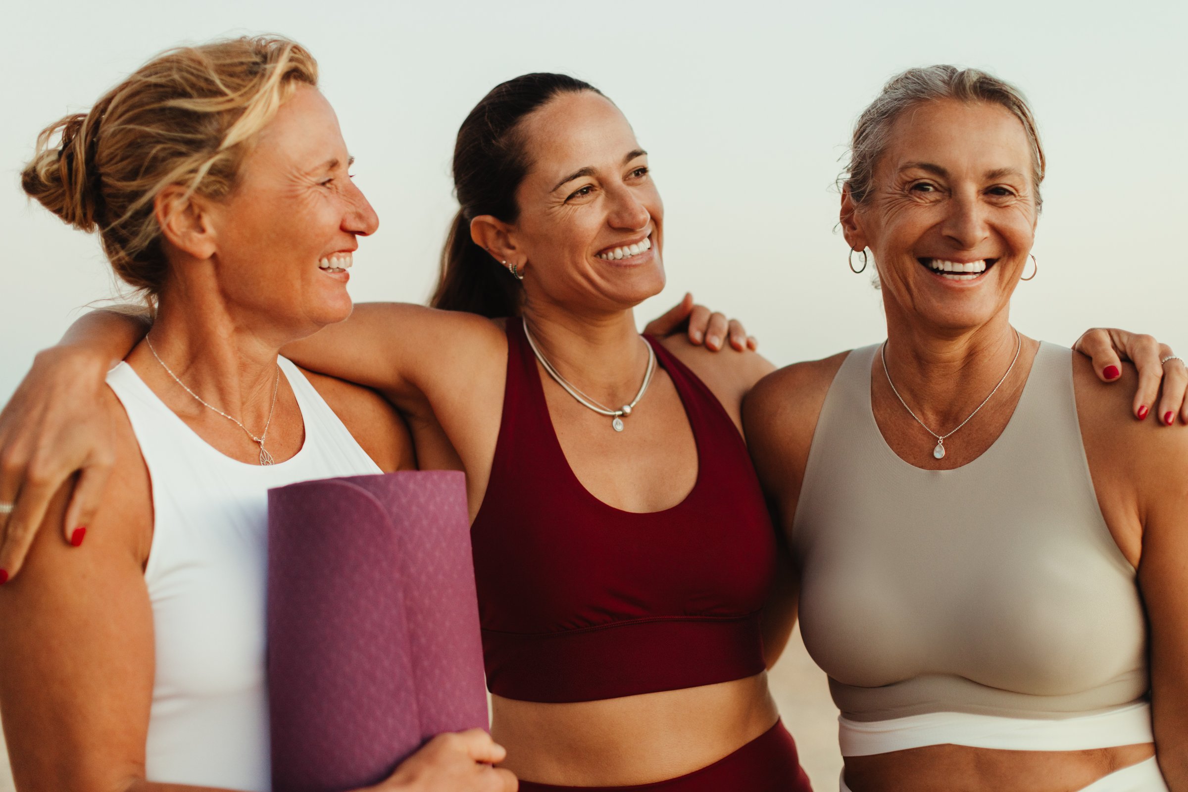 Three mature women share smiles and a bond of friendship while wearing yoga attire outdoors