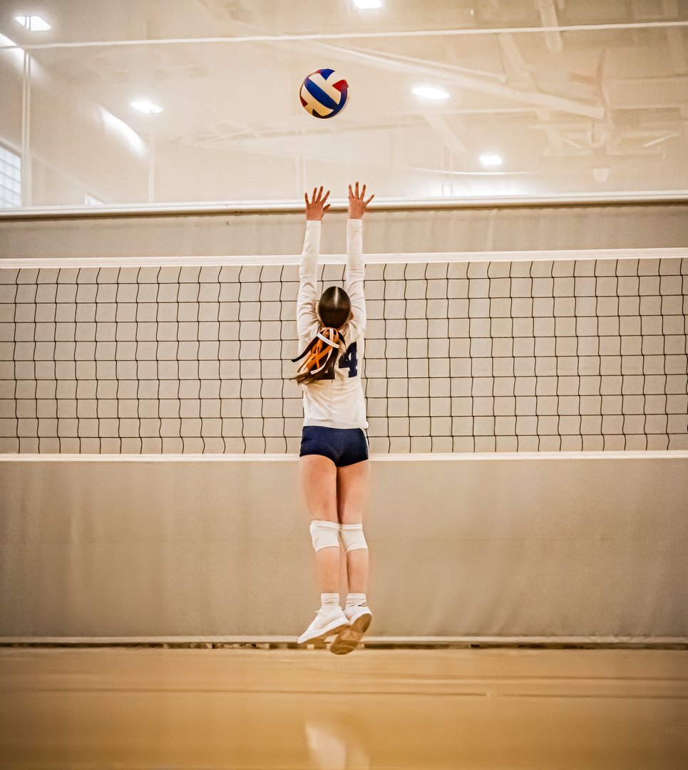 A rear view of a 13 year old female volleyball player in a white uniform going up for a block before a game