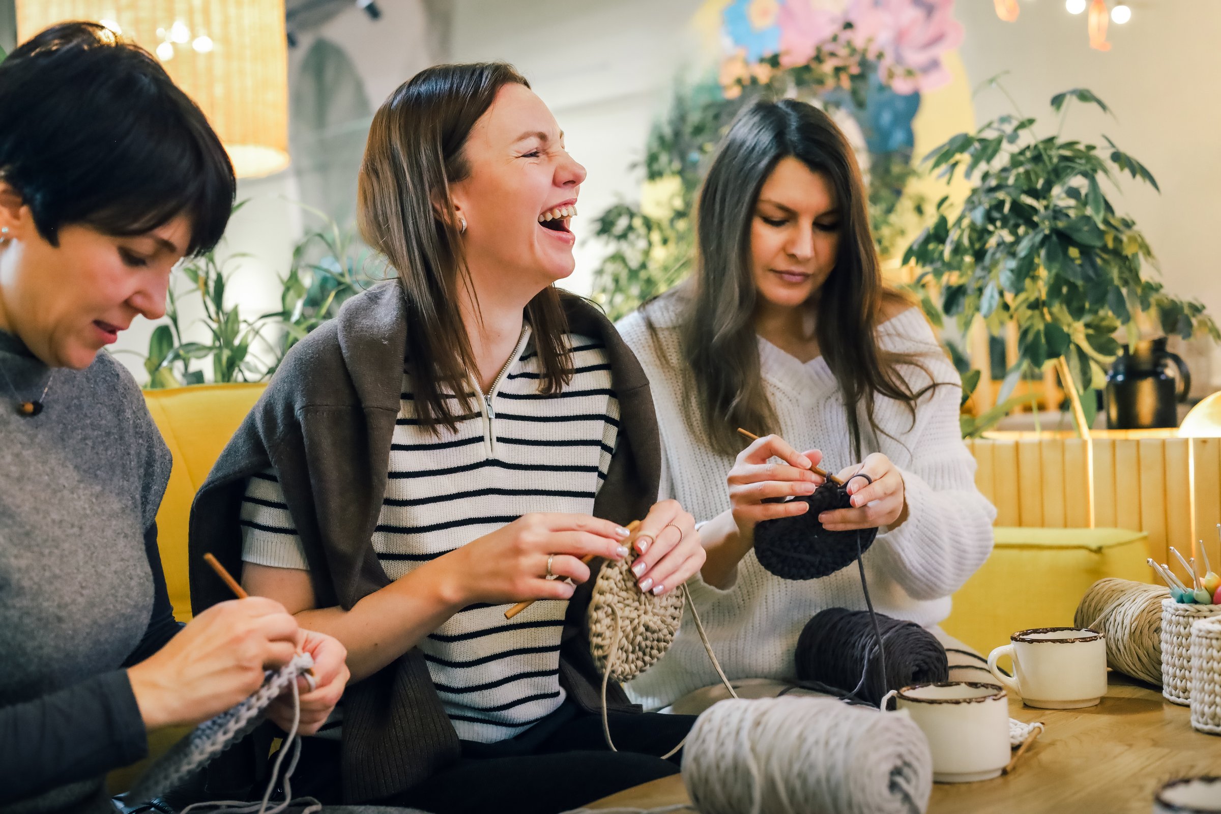 Three women crochet together, sharing laughter and creativity while working on handmade yarn pieces in a warm, inspiring space.