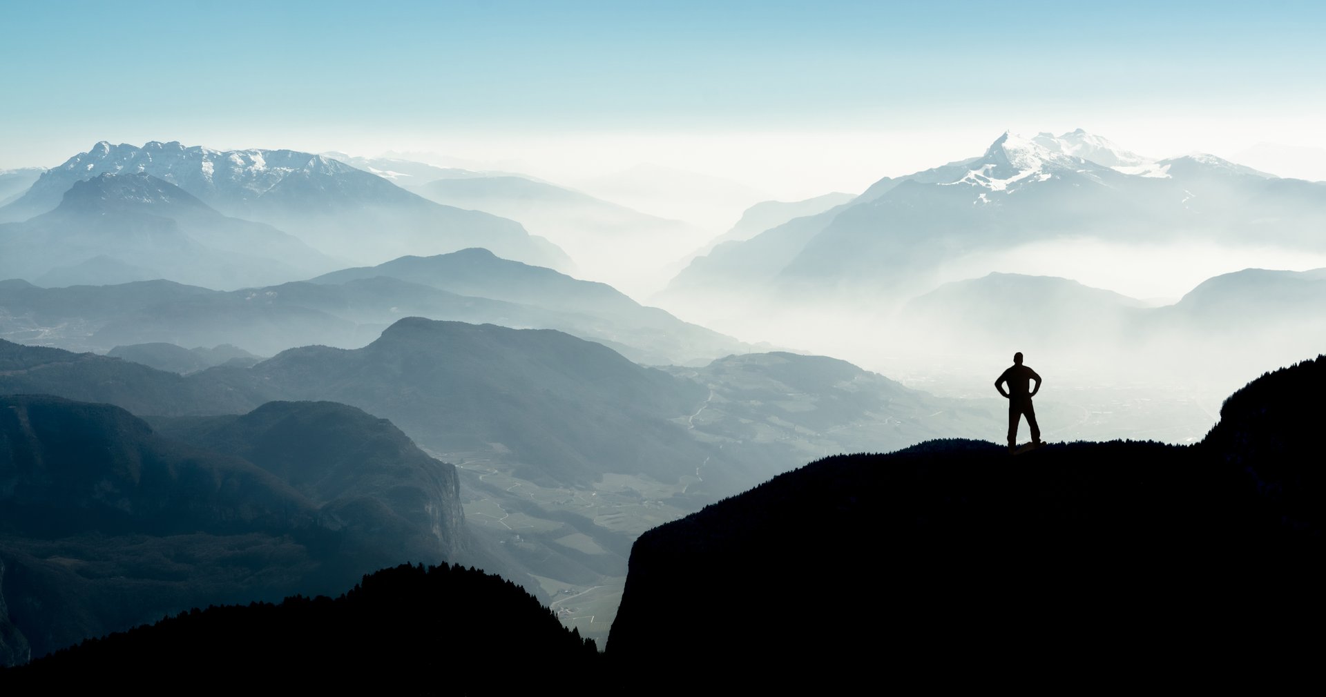 Beautiful view snow covered mountain ranges silhouettes and fog filled valleys with bright back light. South Tyrol, Itay, Alps. Happy winning success man at sunset or sunrise standing relaxed and is happy for having reached mountain top summit goal during hiking travel trek.