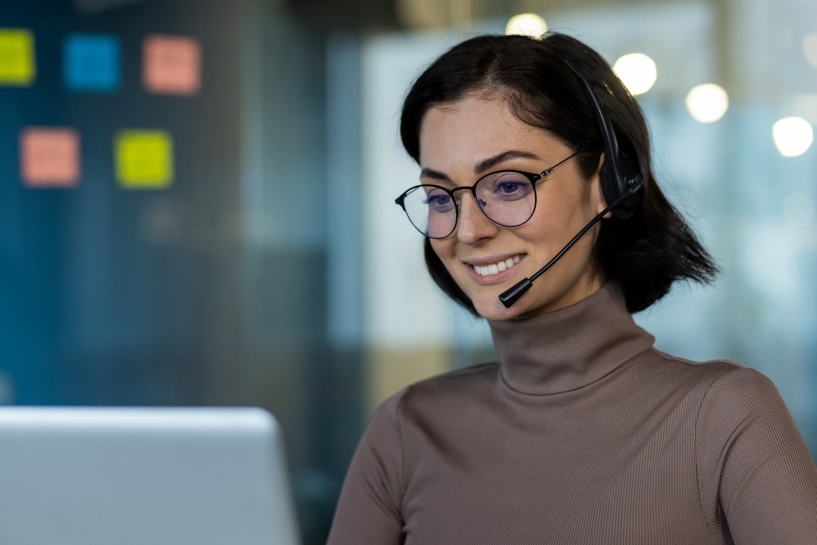 Professional customer service representative using headset while working at laptop. Smiling woman engaging clients over video call, demonstrating communication skills and expertise.