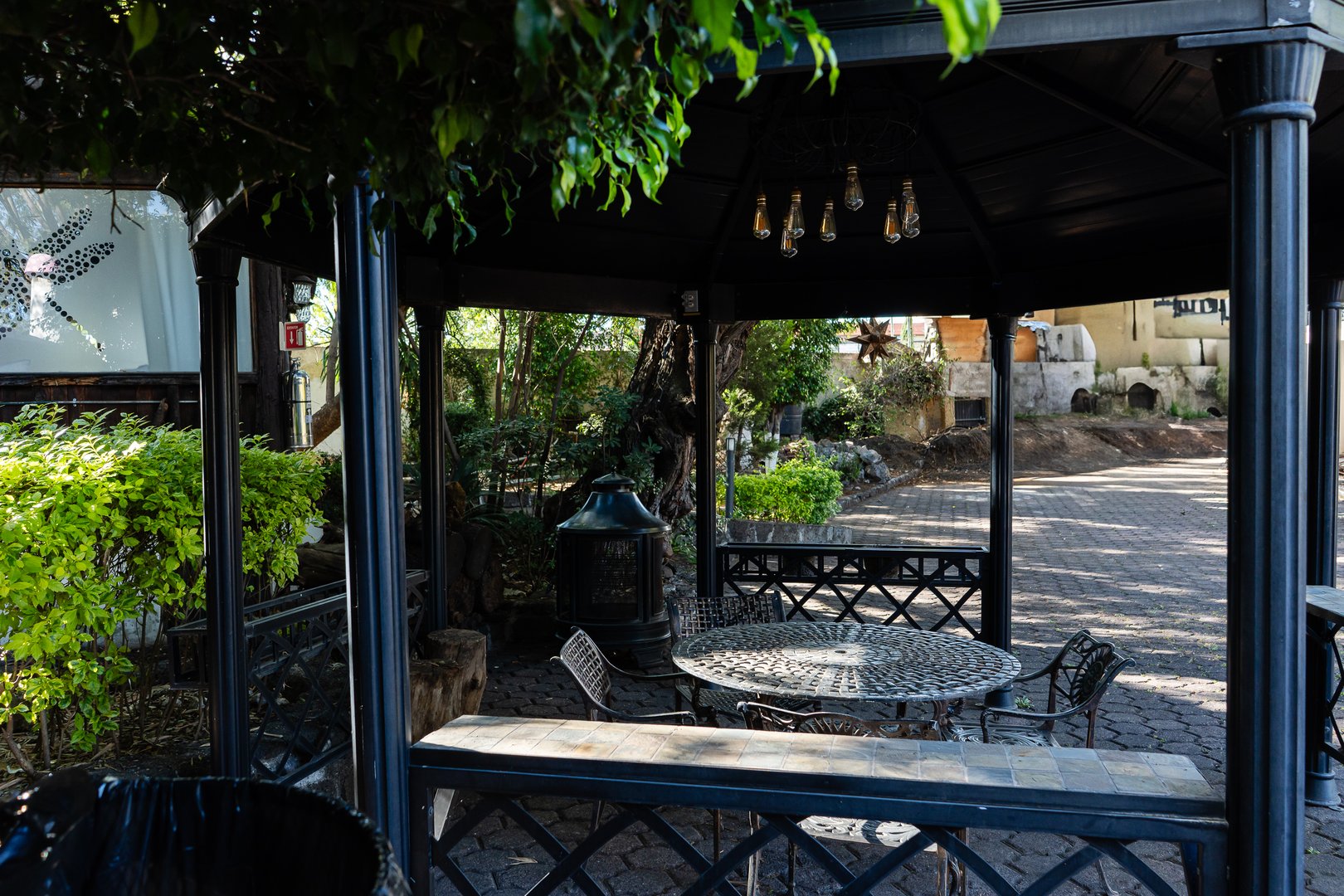 A gazebo with a metal table and chairs underneath, surrounded by lush greenery and a cobblestone path.