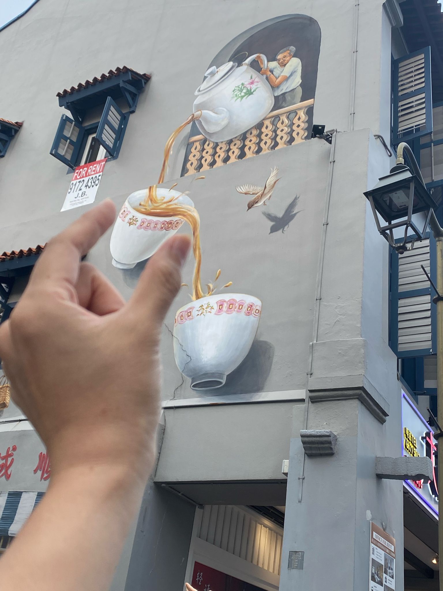 A hand appears to catch tea pouring from a mural of a person pouring tea from a teapot into teacups on a building wall.