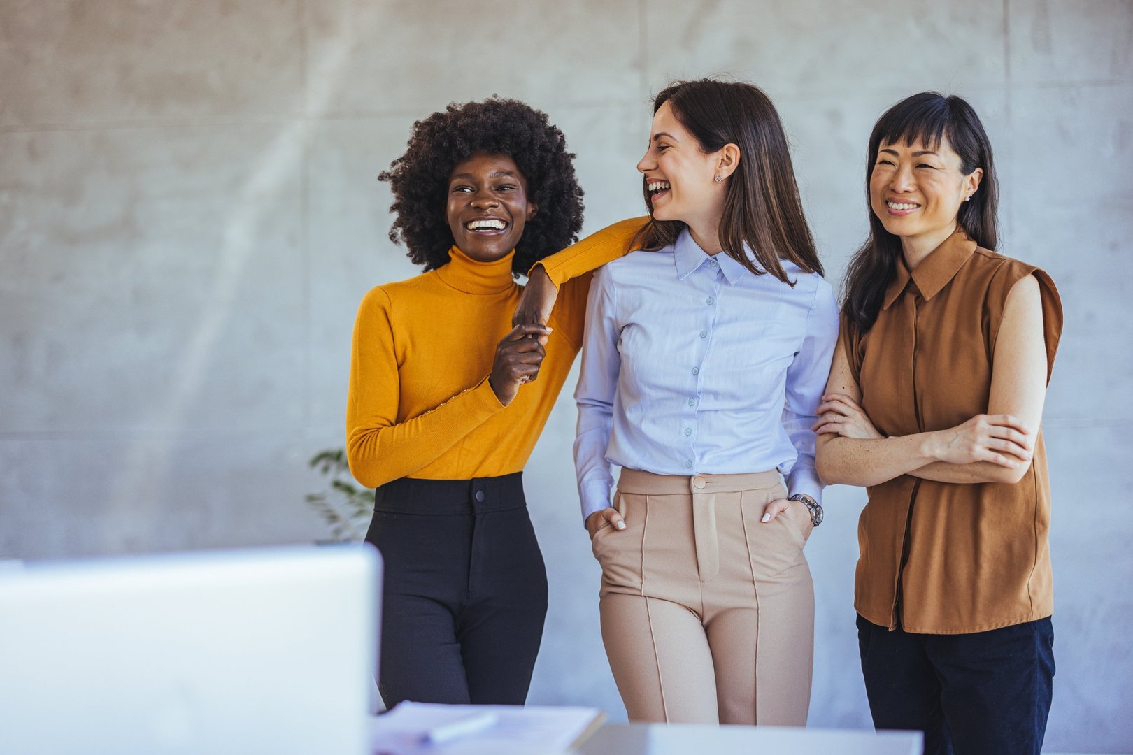 A diverse group of three women smiling and enjoying each other's company in a modern office environment. They exhibit friendship, diversity, and collaboration.