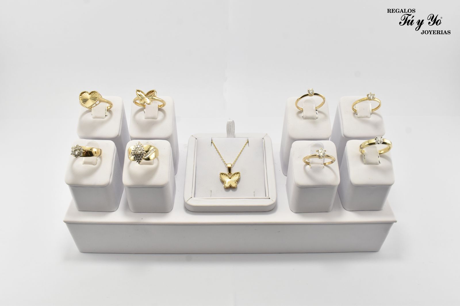 Display of gold rings and a butterfly pendant on stands, arranged on a white platform against a plain background.