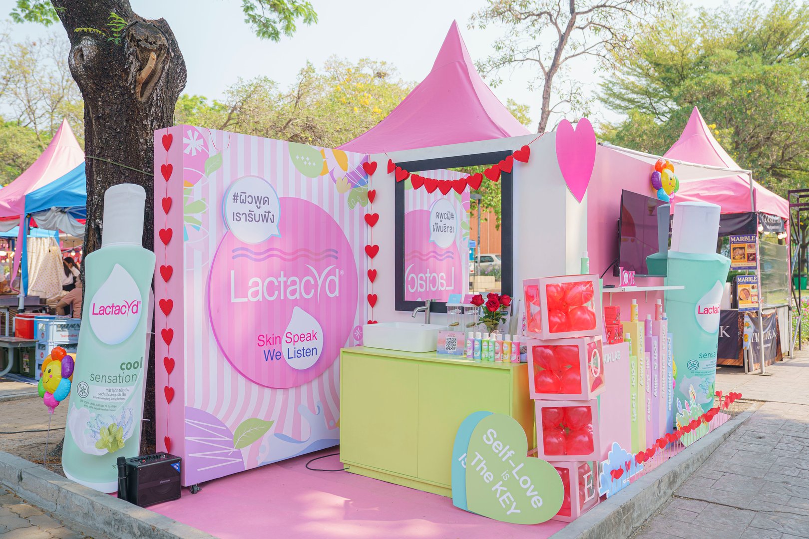 Outdoor booth with Lactacyd branding, featuring product displays and colorful pink tents at a festival setting.