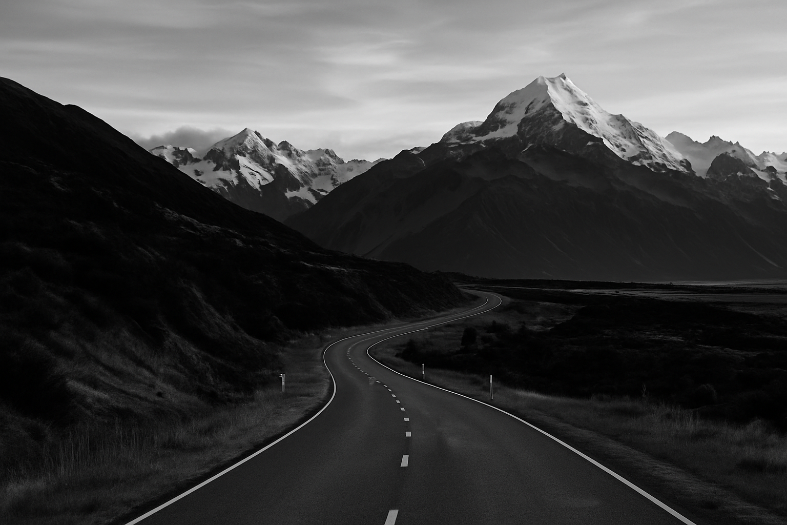 A winding road leading through a mountainous landscape with snow-capped peaks under a cloudy sky.
