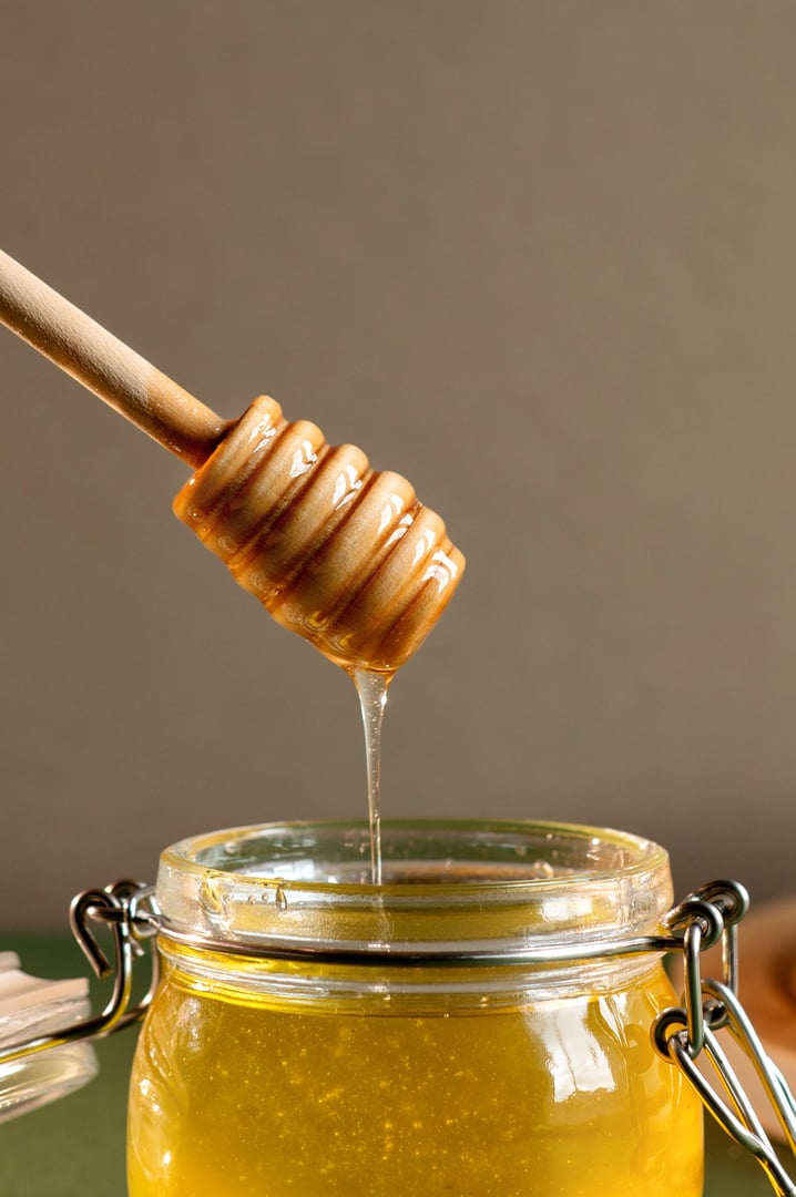 Closed up a glass jar filled with golden honey and a wooden dipper dripping honey against a neutral background.