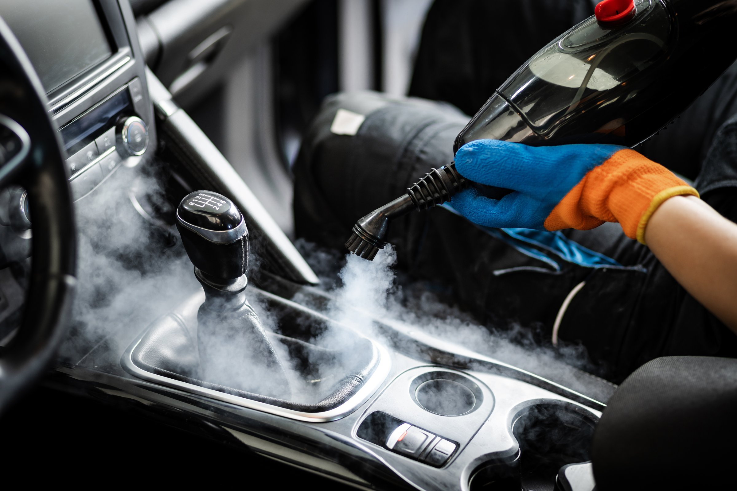 A person is using a steam cleaner inside a car, focusing on the gear shift area, releasing steam to eliminate stains and disinfect surfaces effectively.