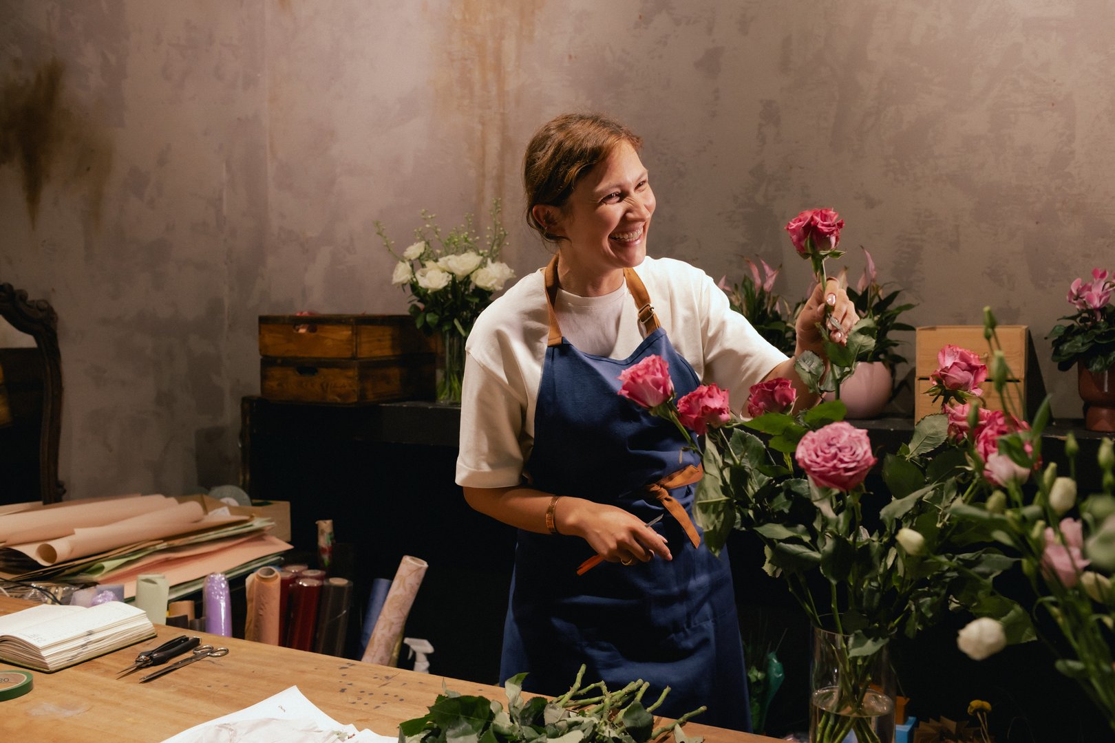 Middle-aged female florist in apron arranging fresh flowers for bouquet in the flower shop. Small local business. Part of a series. Soft focus