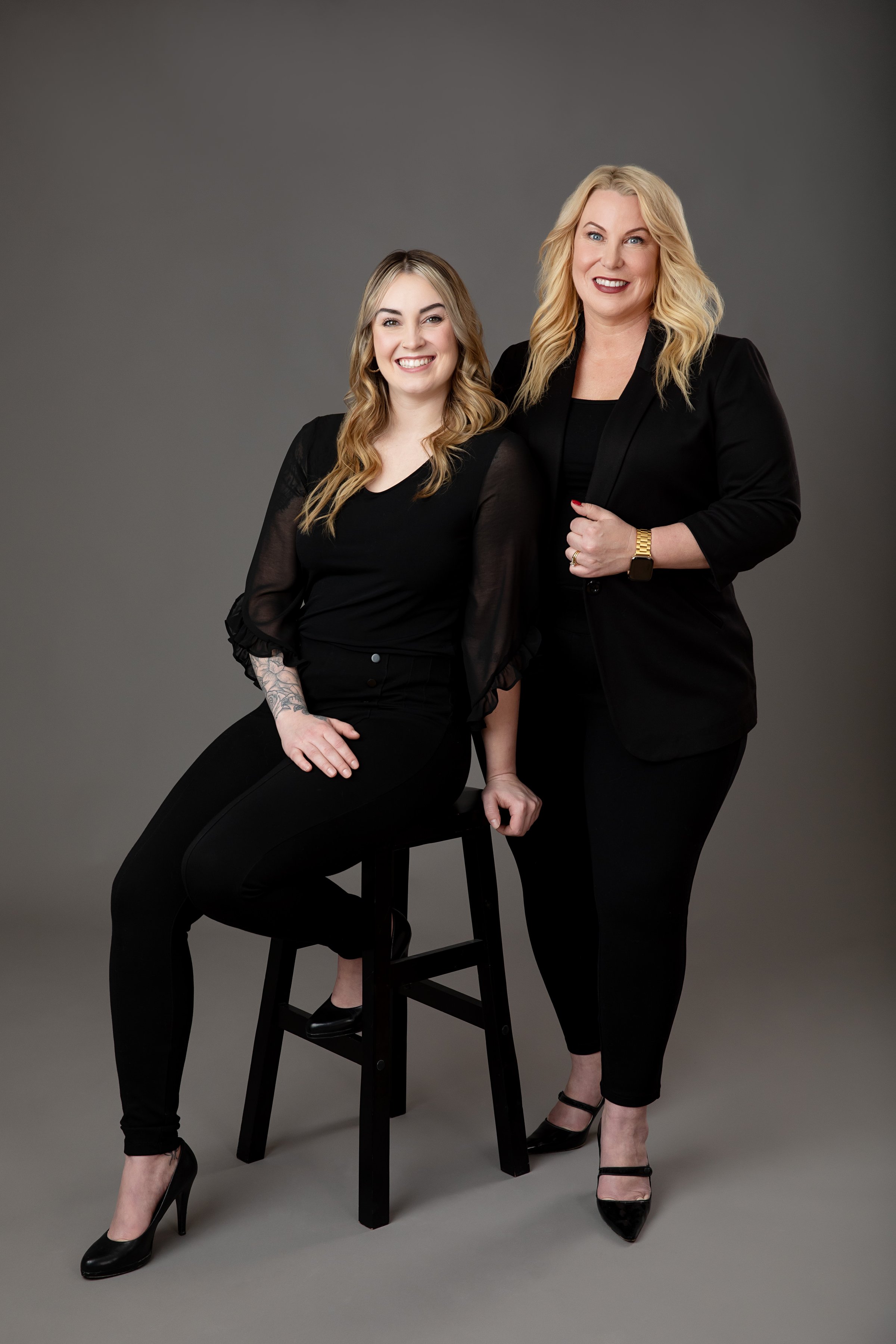 Two women in black attire, one seated on a stool and the other standing, both smiling in a studio setting.