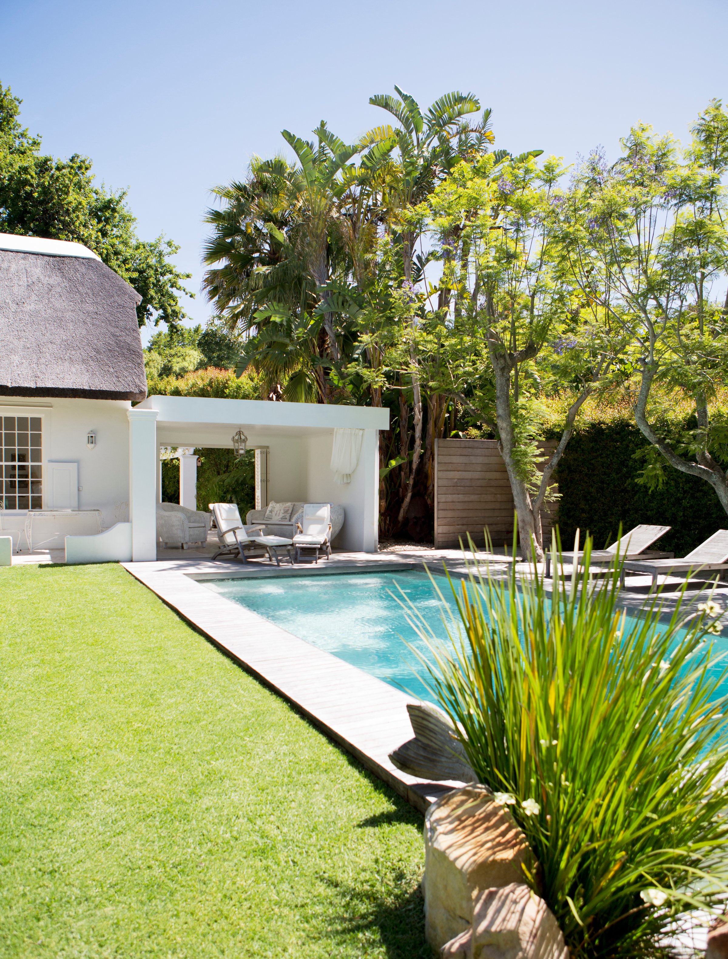 A serene backyard with a pool, lounge chairs, lush greenery, and a white thatched-roof cabana under a clear blue sky.