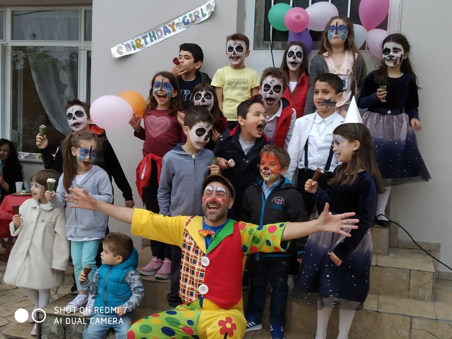 A group of children with face paint and a clown pose for a photo at a birthday party, with balloons and a banner in the background.