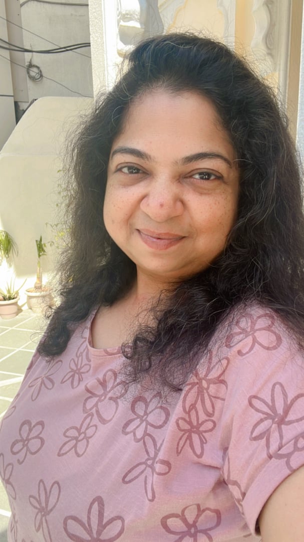 A woman with curly hair smiling, wearing a floral pink top, standing outdoors with a sunlit background.