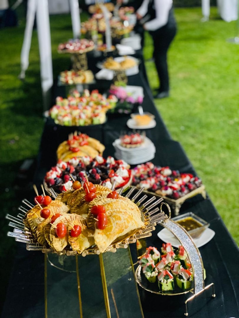 A buffet table outdoors with various appetizers including pastries, fruit skewers, and rolls. A server stands in the background.