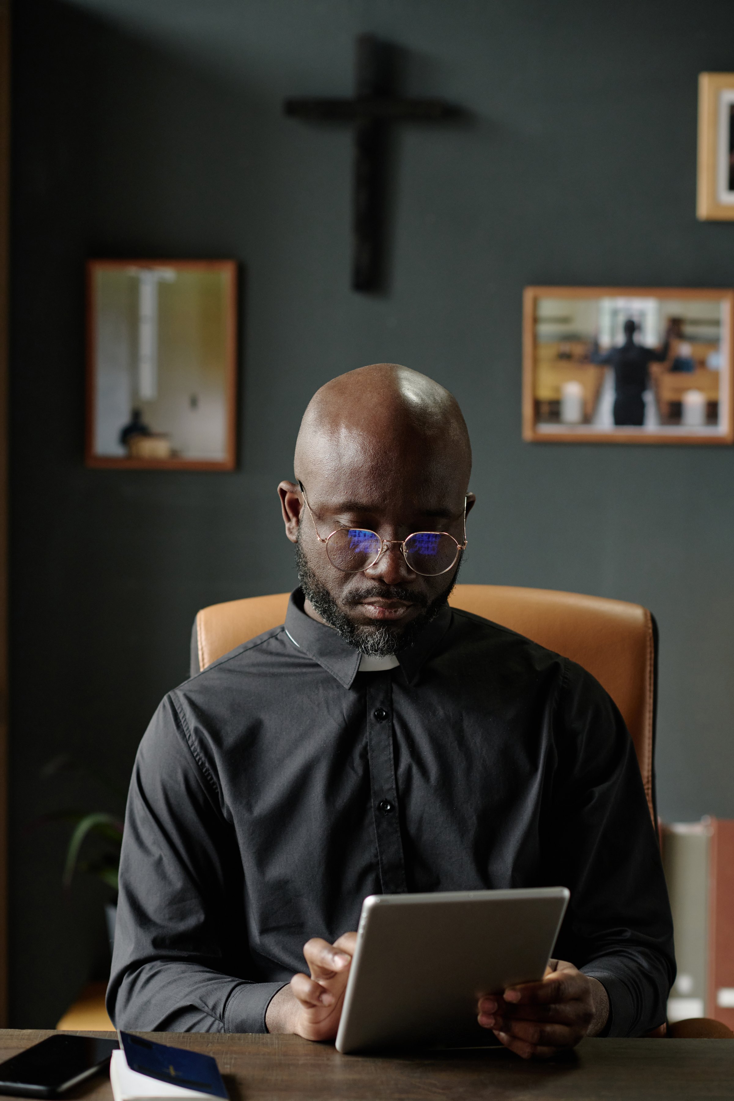 Vertical shot of African American pastor using tablet to check email while sitting at table in office