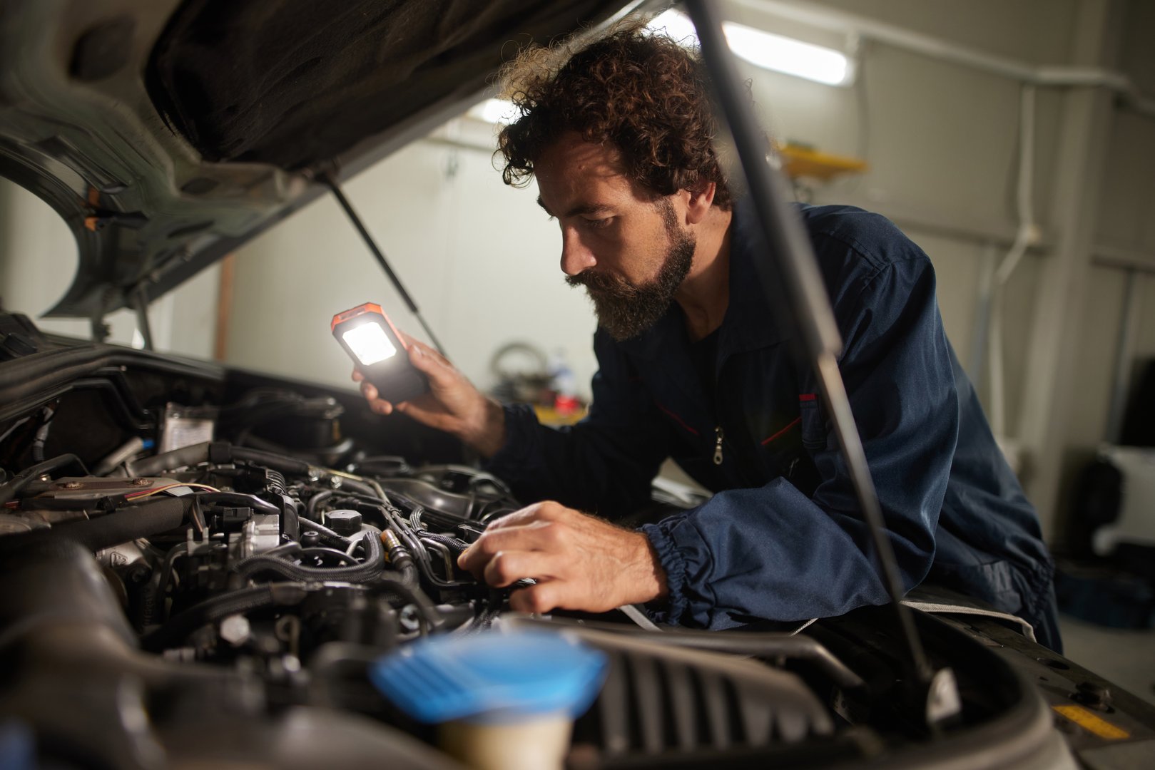 Mid adult car mechanic inspecting car engine with flashlight in a garage
