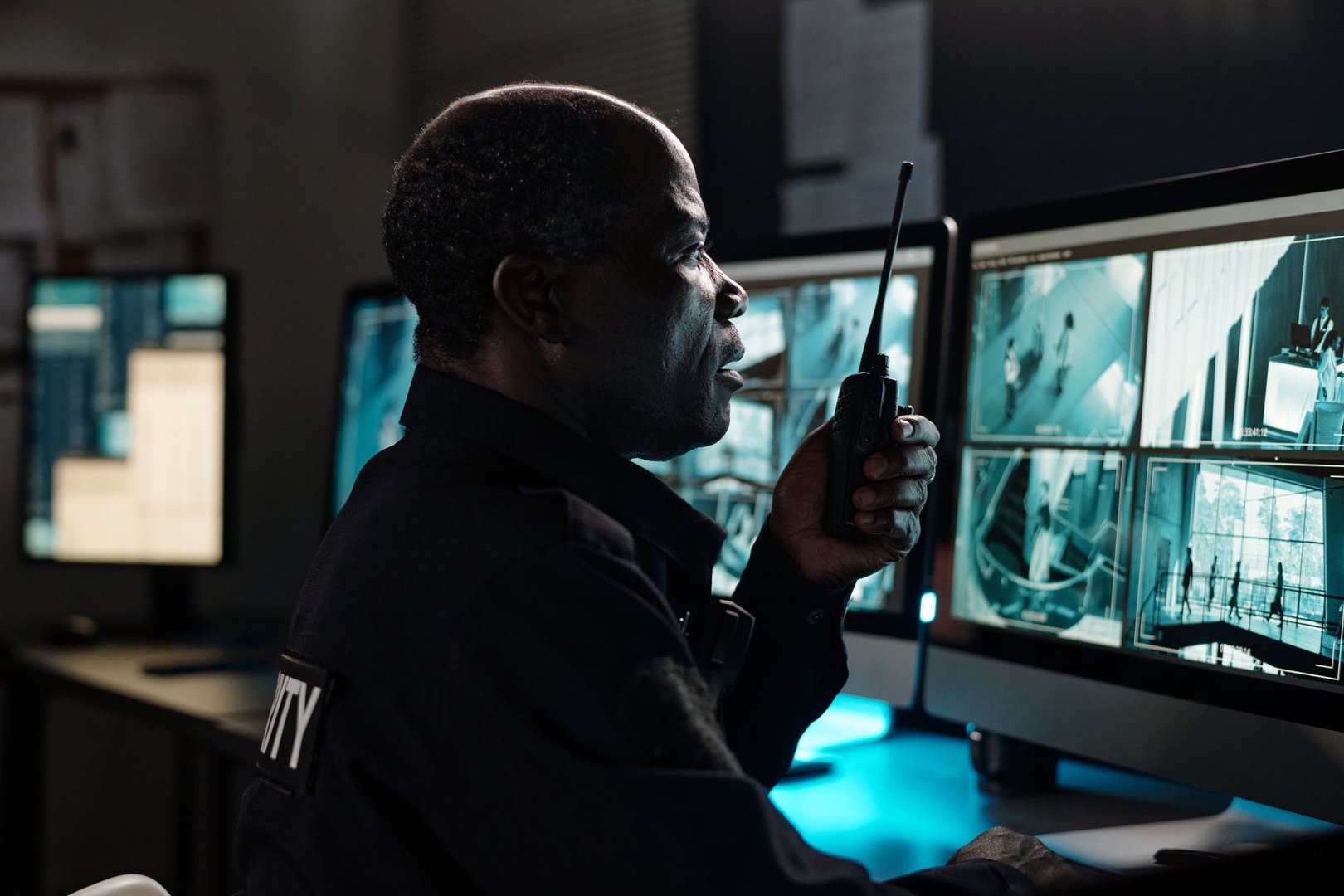 Side view of mature African American male guard with walkie-talkie in hand sitting in front of desktop computer monitors
