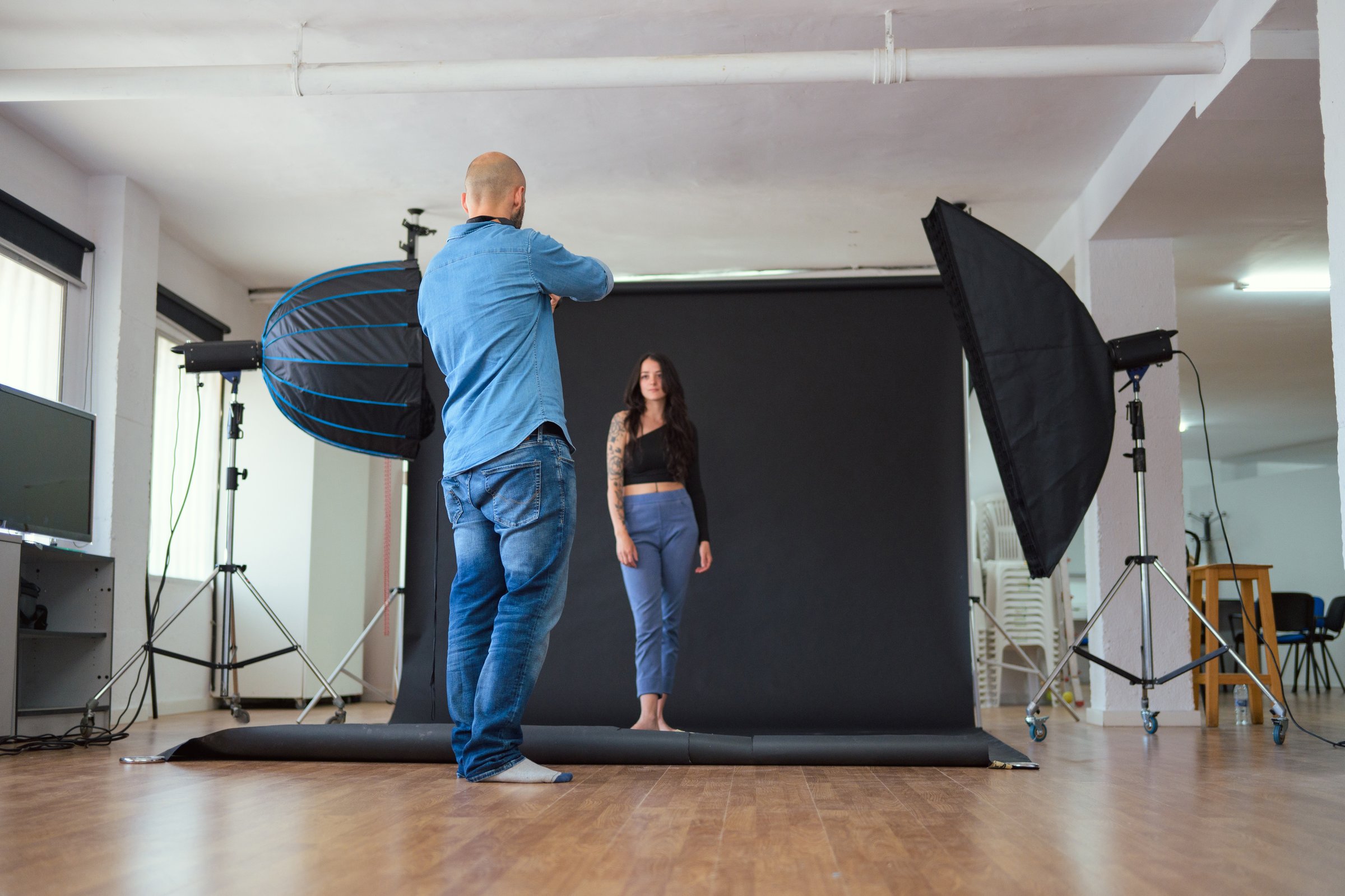A photographer shoots a female model in a studio. The model is standing, wearing a black top and blue jeans. The photographer is holding a camera. The background is a black backdrop a with softbox