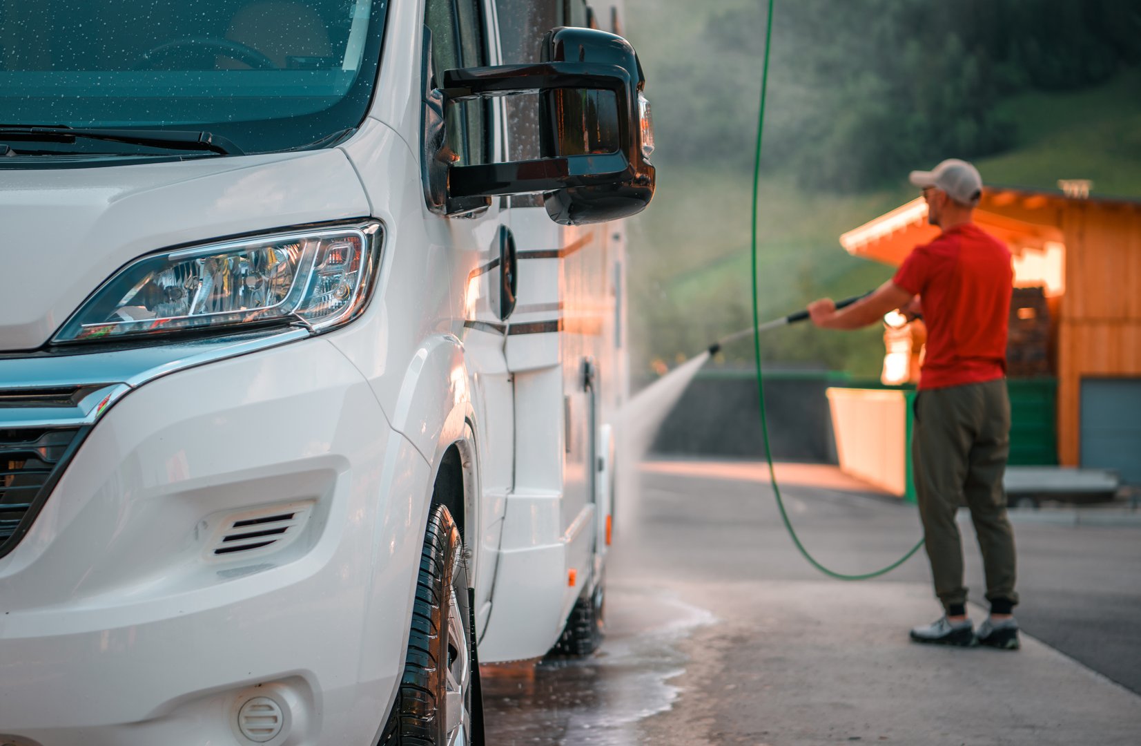 A man is spraying water on a white camper van, cleaning it thoroughly at a service station with lush greenery in the background.