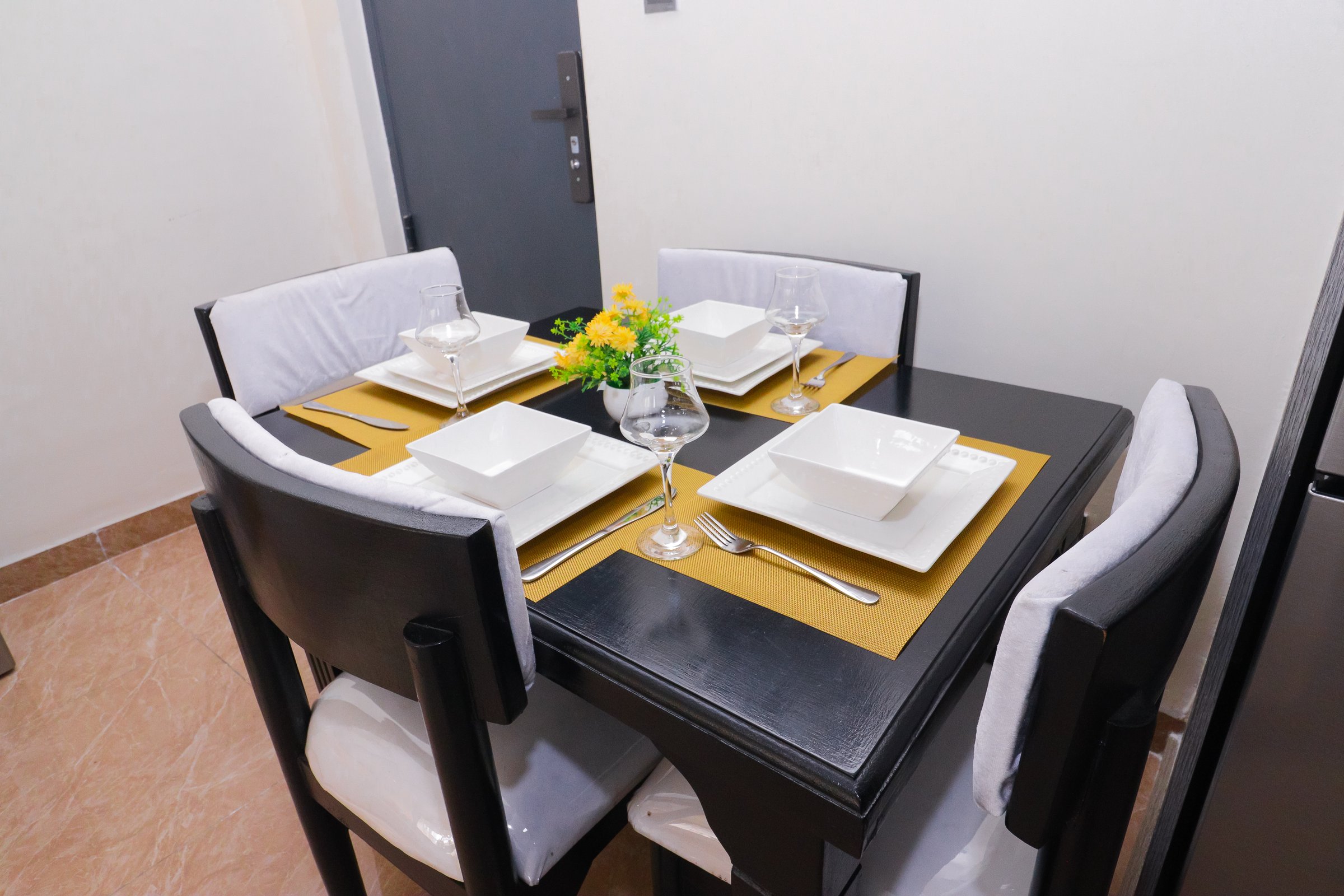 Elegant dining table set for four with square plates, wine glasses, and yellow placemats, featuring a flower centerpiece.