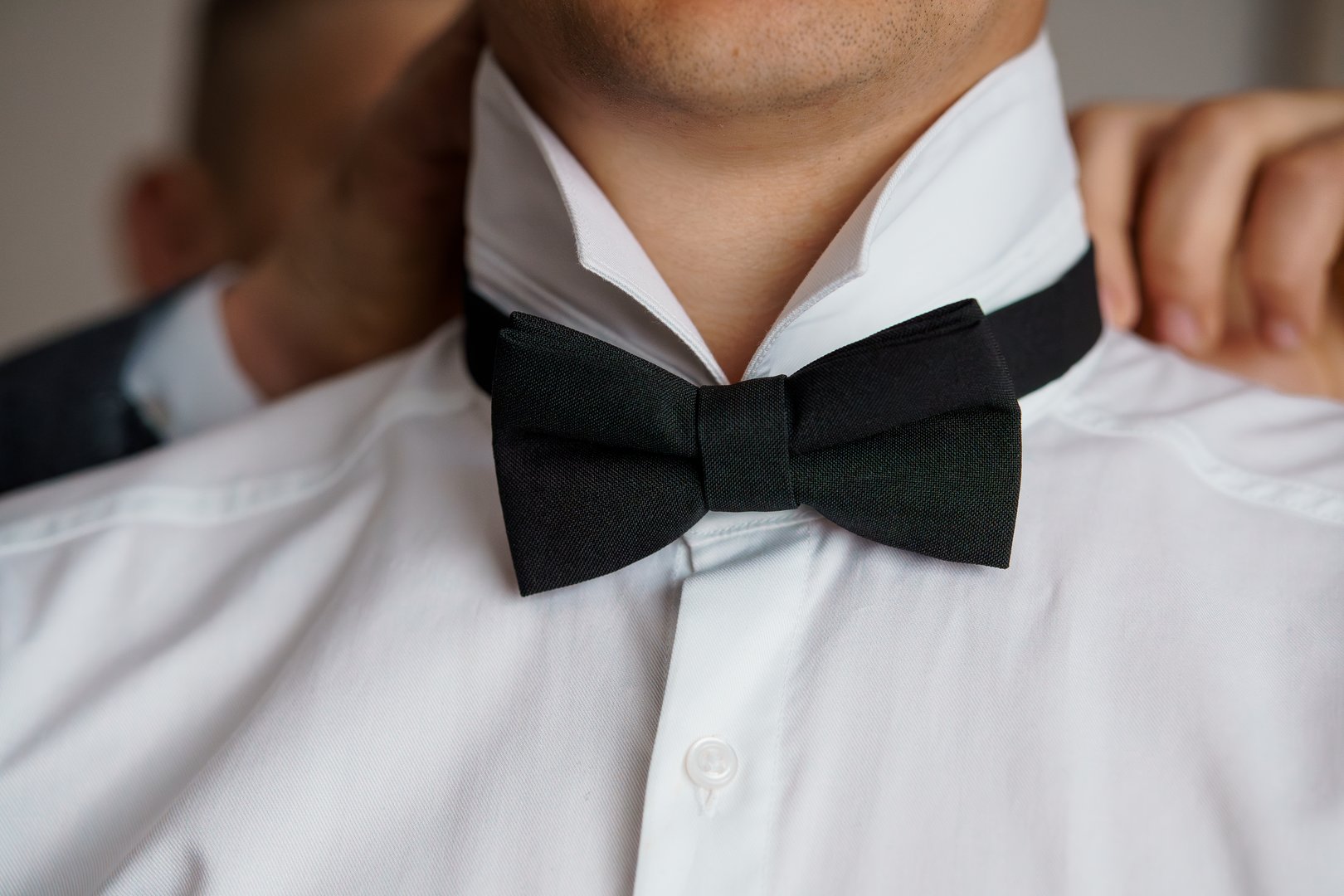 A close-up portrait depicts a groom getting ready, someone adjusting his black bow tie on a crisp white shirt, symbolizing elegance, preparation, and weddings