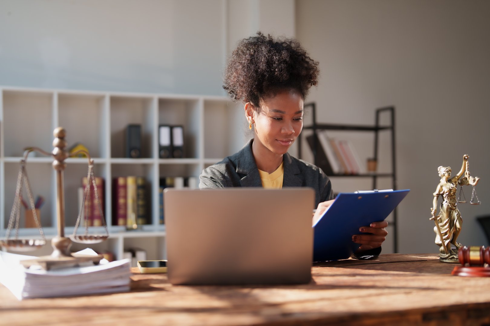 Female lawyer sitting at her desk, concentrating while writing on a clipboard, with a laptop, scales of justice, and a lady justice statue nearby