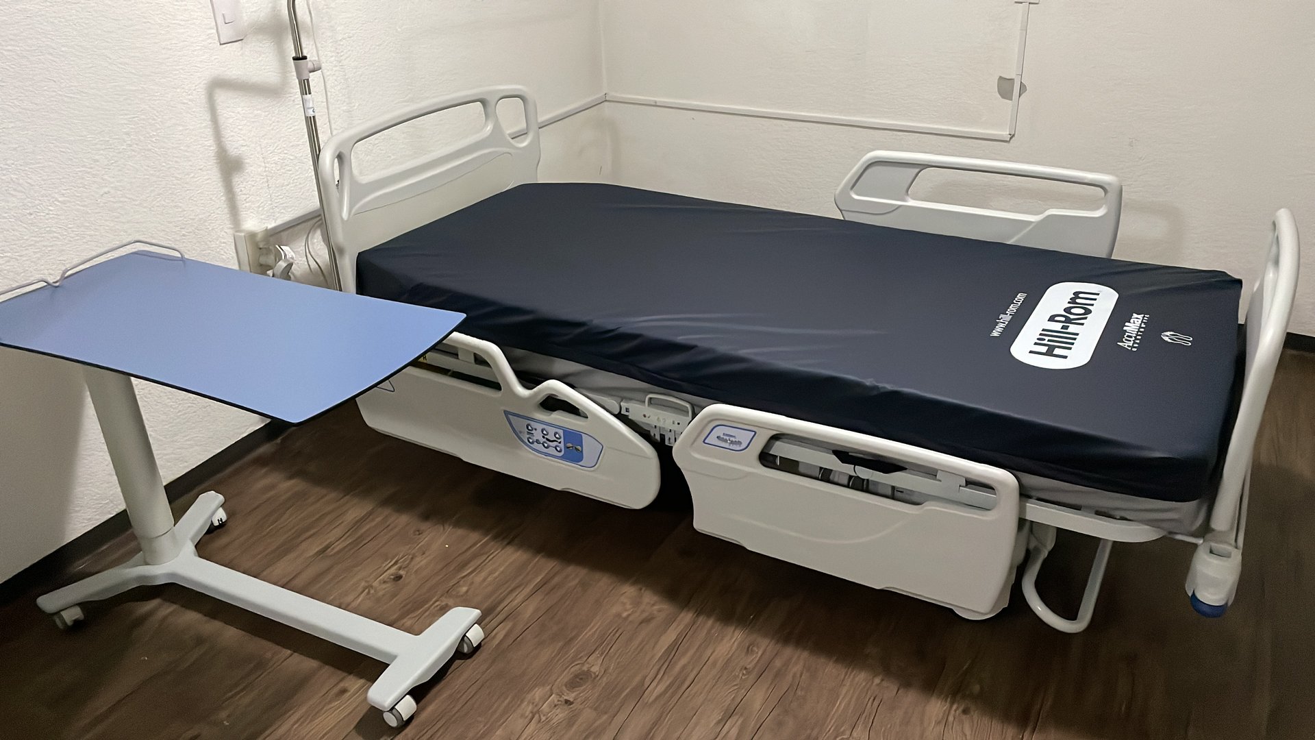 A hospital bed with a dark mattress and side rails, next to a blue adjustable table, in a room with wooden flooring.