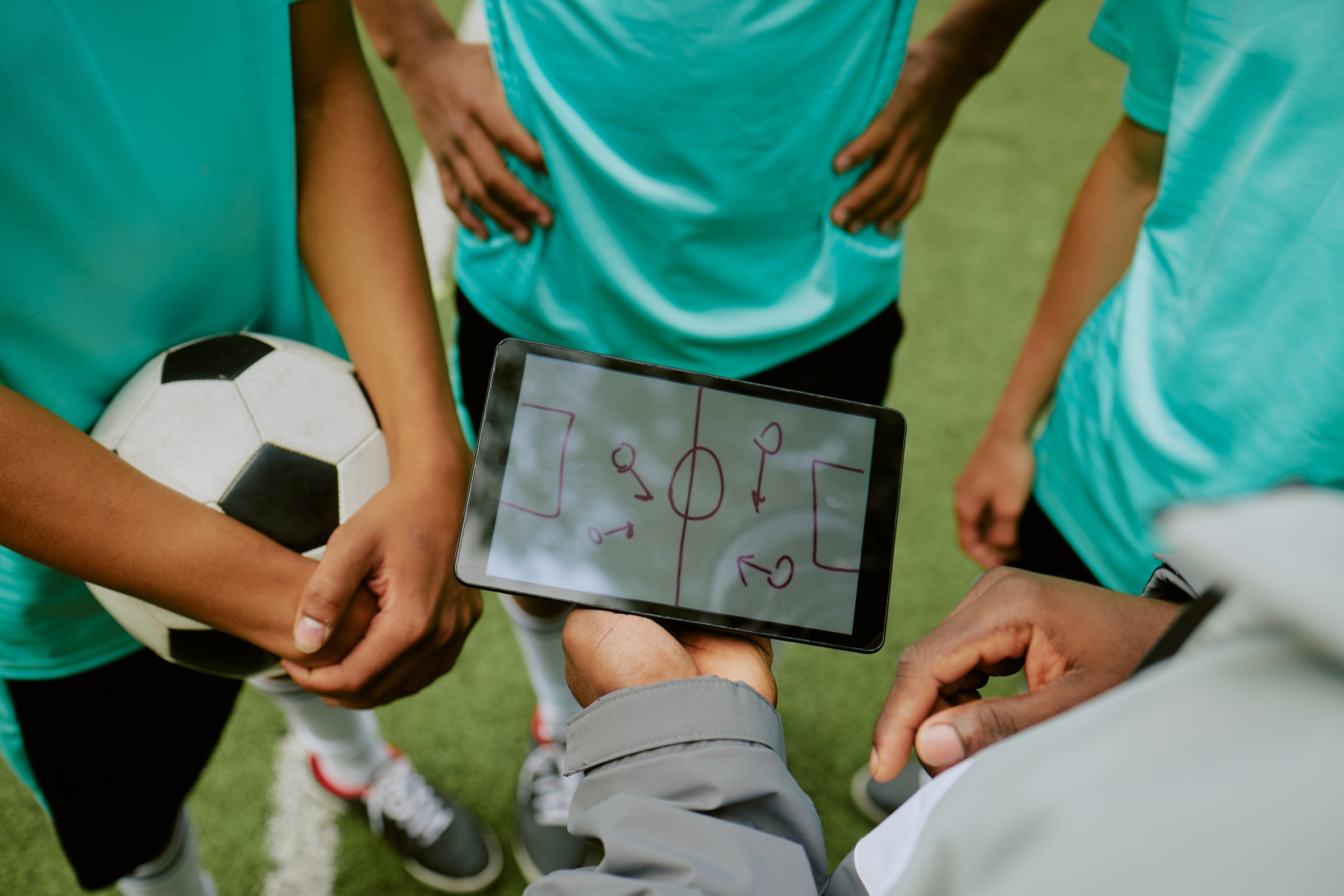 Group of teenage boys standing on soccer field listening to coach holding tablet displaying tactical diagram, one teenager holding soccer ball during team discussion