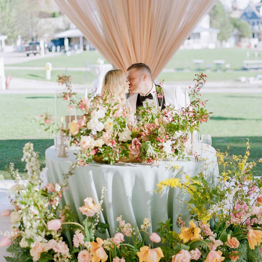 Couple kissing at a flower-adorned table under a canopy in an outdoor setting.