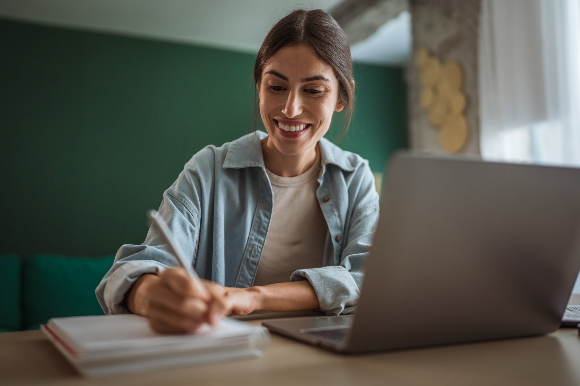 Young woman smiling while taking notes in a notebook, working on a laptop at home. Enjoying the flexibility of remote work and the benefits of online education
