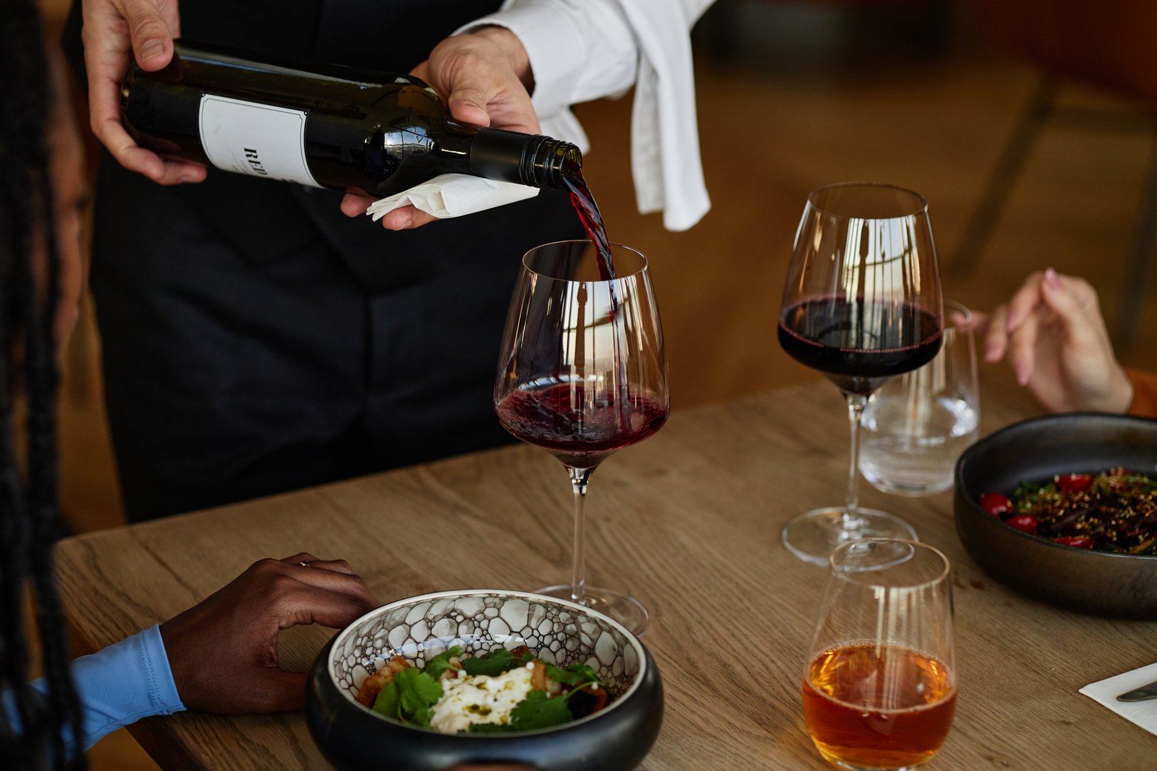 Man pouring red wine into glass for diverse group of young adults sitting at restaurant table with food and drinks