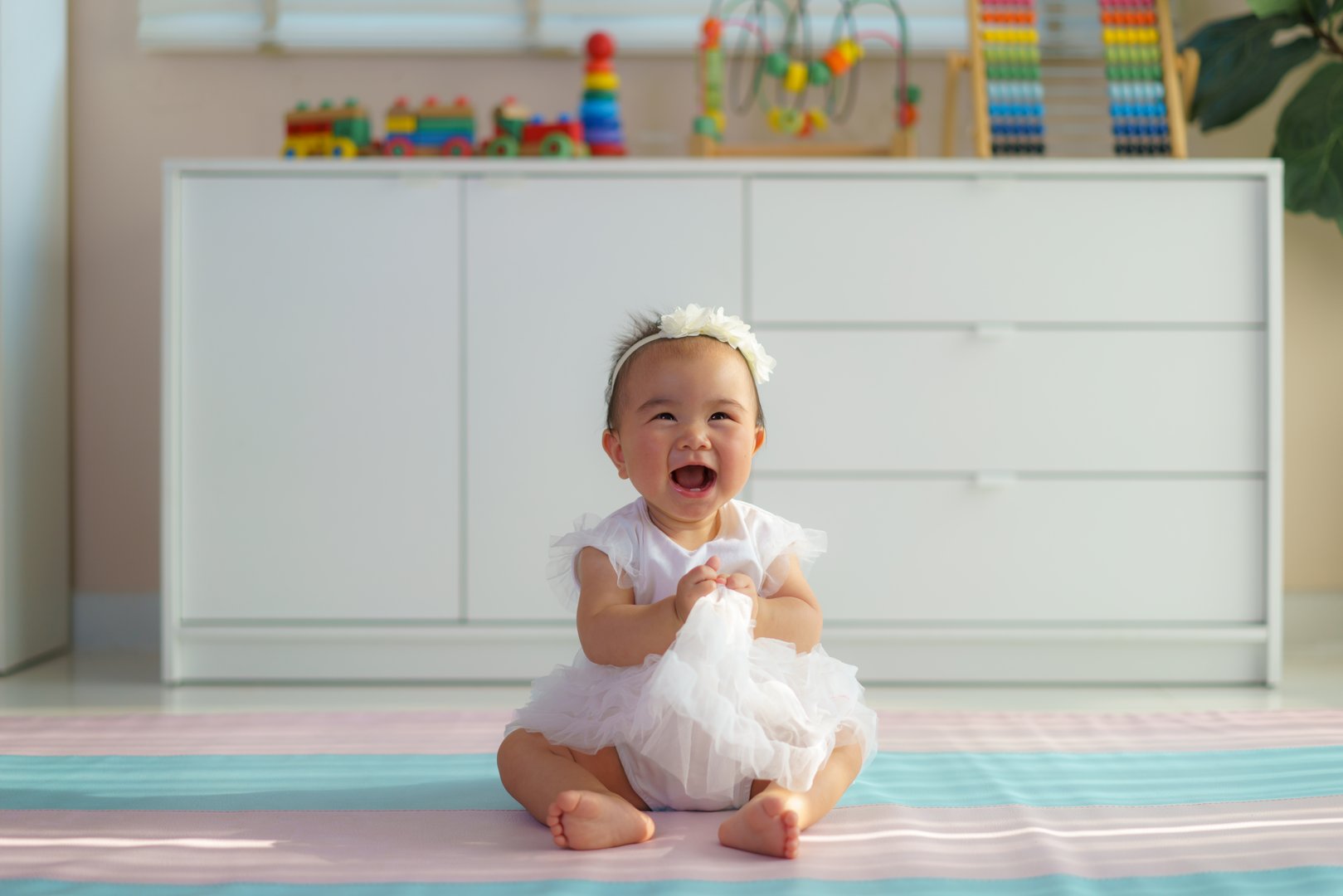 joyful baby girl in a frilly white dress laughs while sitting on a colorful playmat, showcasing innocence, happiness, and warm indoor play.