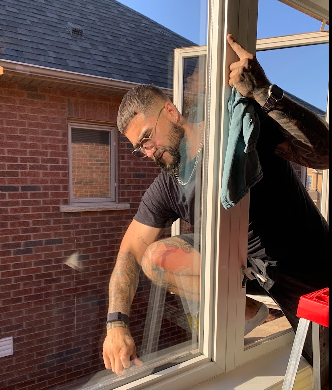 Man cleaning an open window with a cloth, wearing glasses and a smartwatch, kneeling on a step ladder under bright sunlight.