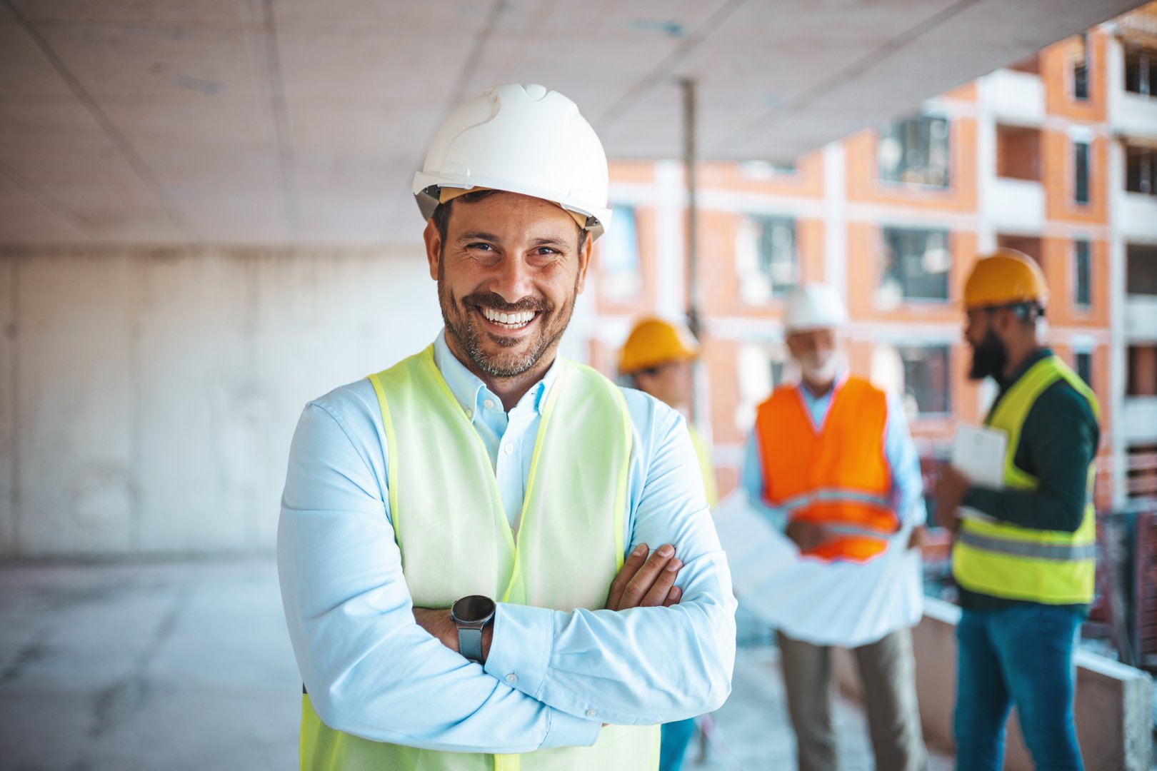 Portrait of man architect at building site with folded arms looking at camera. Confident construction manager in formal clothing wearing white hardhat. Successful mature civil engineer at construction site with copy space.