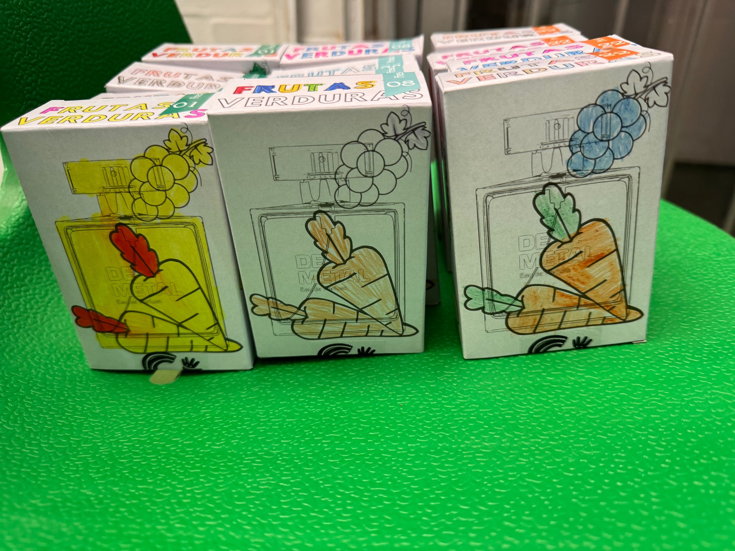 Colorful perfume boxes featuring fruit and vegetable designs atop each bottle, arranged on a bright green surface.
