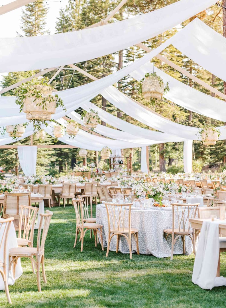 Outdoor wedding reception with white draped ceiling, wooden chairs, and elegantly decorated round tables on a grassy lawn.