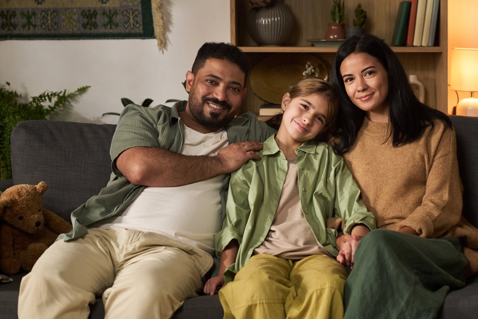Hispanic family sitting on cozy couch smiling warmly at camera, with child nestled between parents and dimly lit home decor adding warmth and comfort