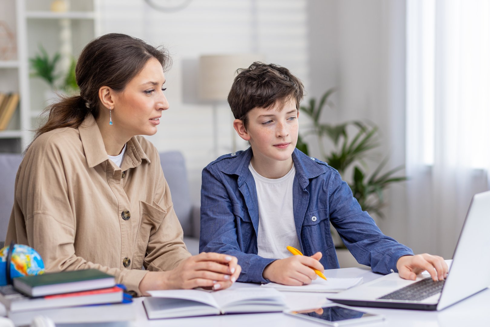 Mother and son studying together sitting at desk at home in living room, boy and woman tutor teaching at home, doing homework together.
