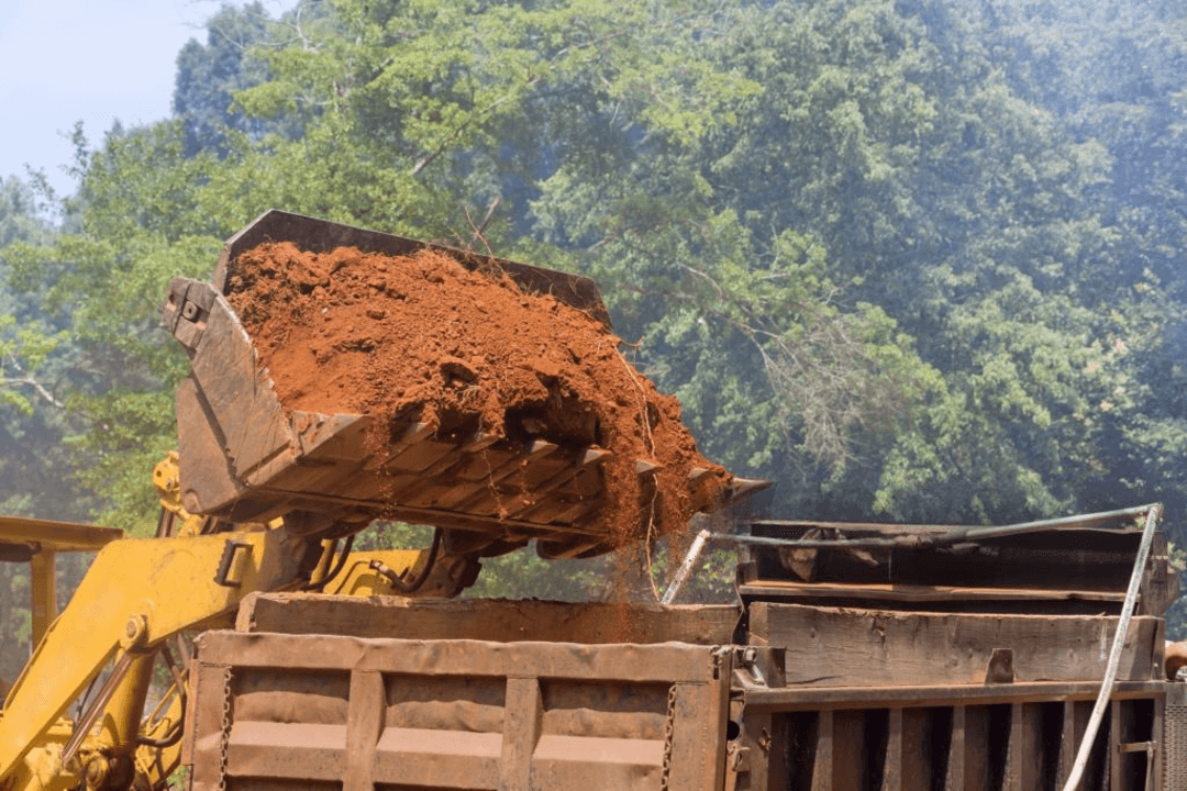 A backhoe loader pours red soil into a truck bed, surrounded by trees in the background.