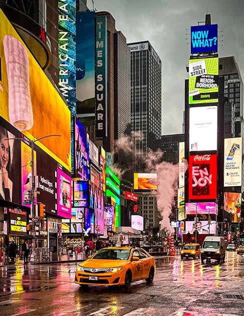 Colorful view of Times Square with bright billboards, steam rising, and yellow taxis on a wet street.