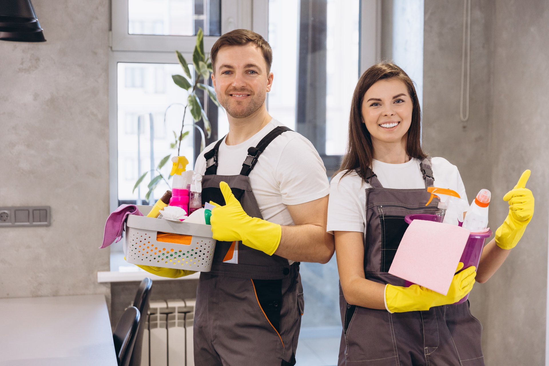 Professional cleaners wearing uniform and yellow gloves holding cleaning products and showing thumbs up