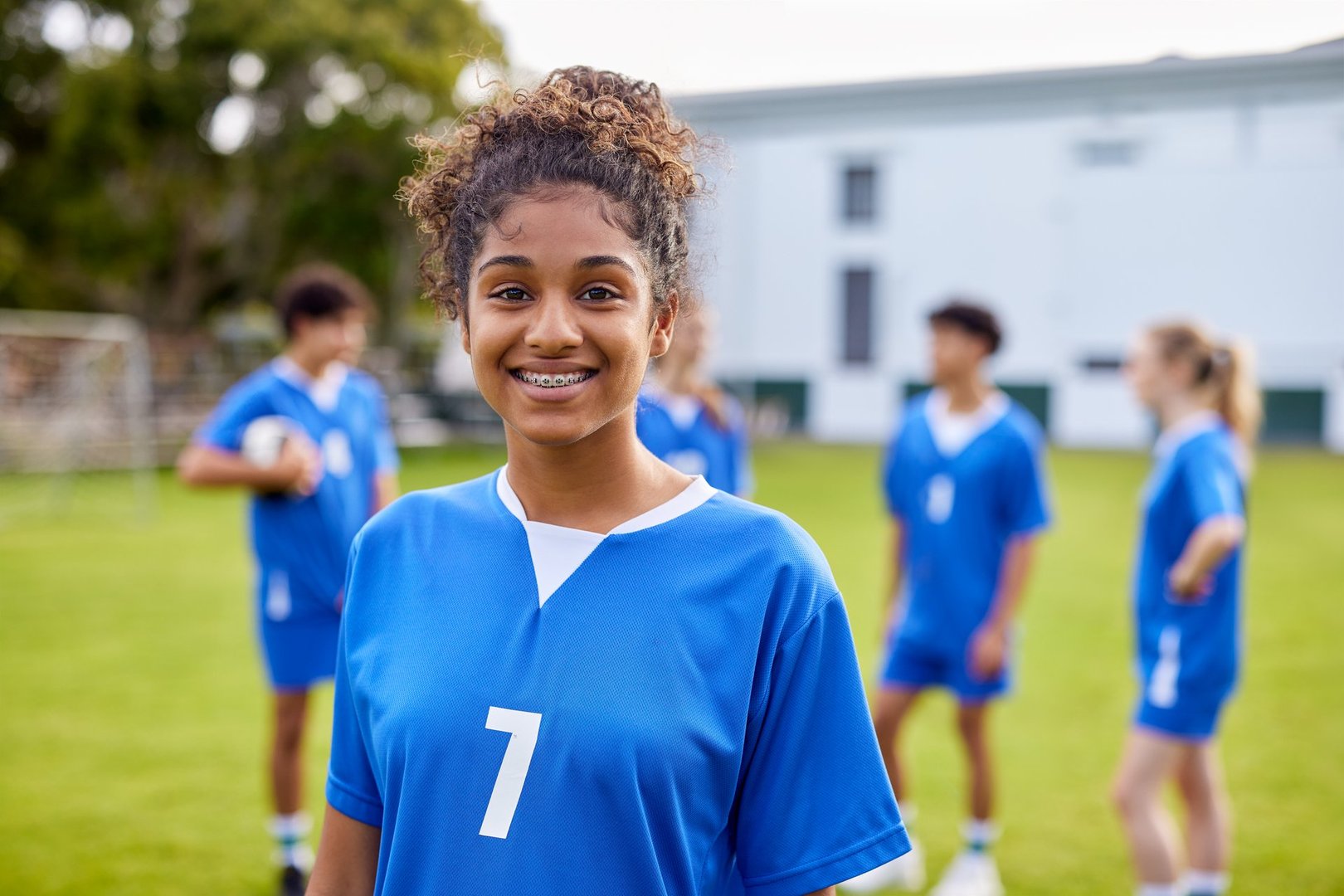 Smiling black teenager wearing blue sports uniform stand confidently on grassy field while looking at camera with copy space. Portrait of confident student girl athlete smiling with dental device at school gymnasium. Fitness girl enjoy sports time in school during gymnastics class.