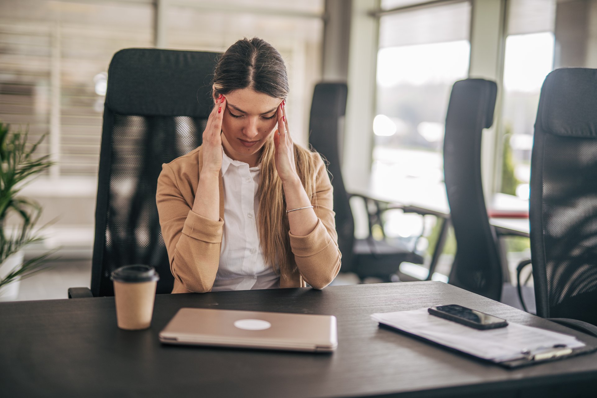 Young businesswoman sitting at her desk with a laptop, documents and smartphone, suffering from a headache and touching her hands with her hands