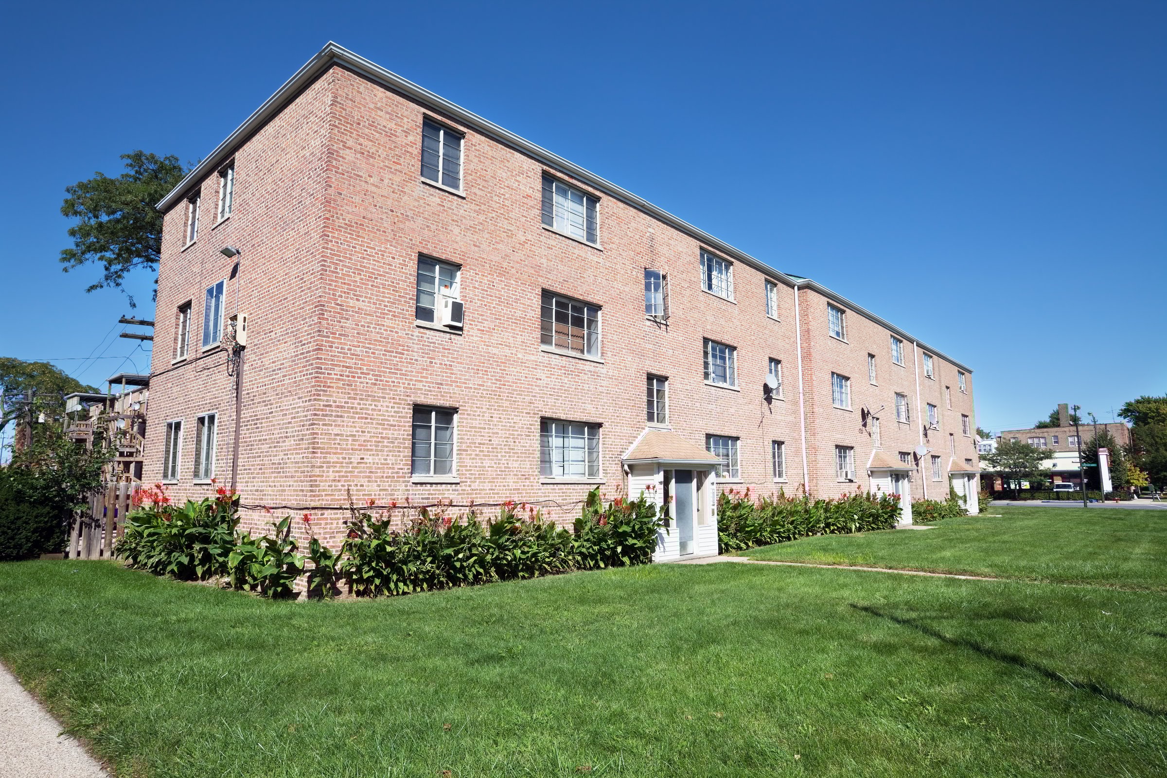 Three-story brick apartment building with multiple units, white entrance doors, and landscaped lawn with flowering plants