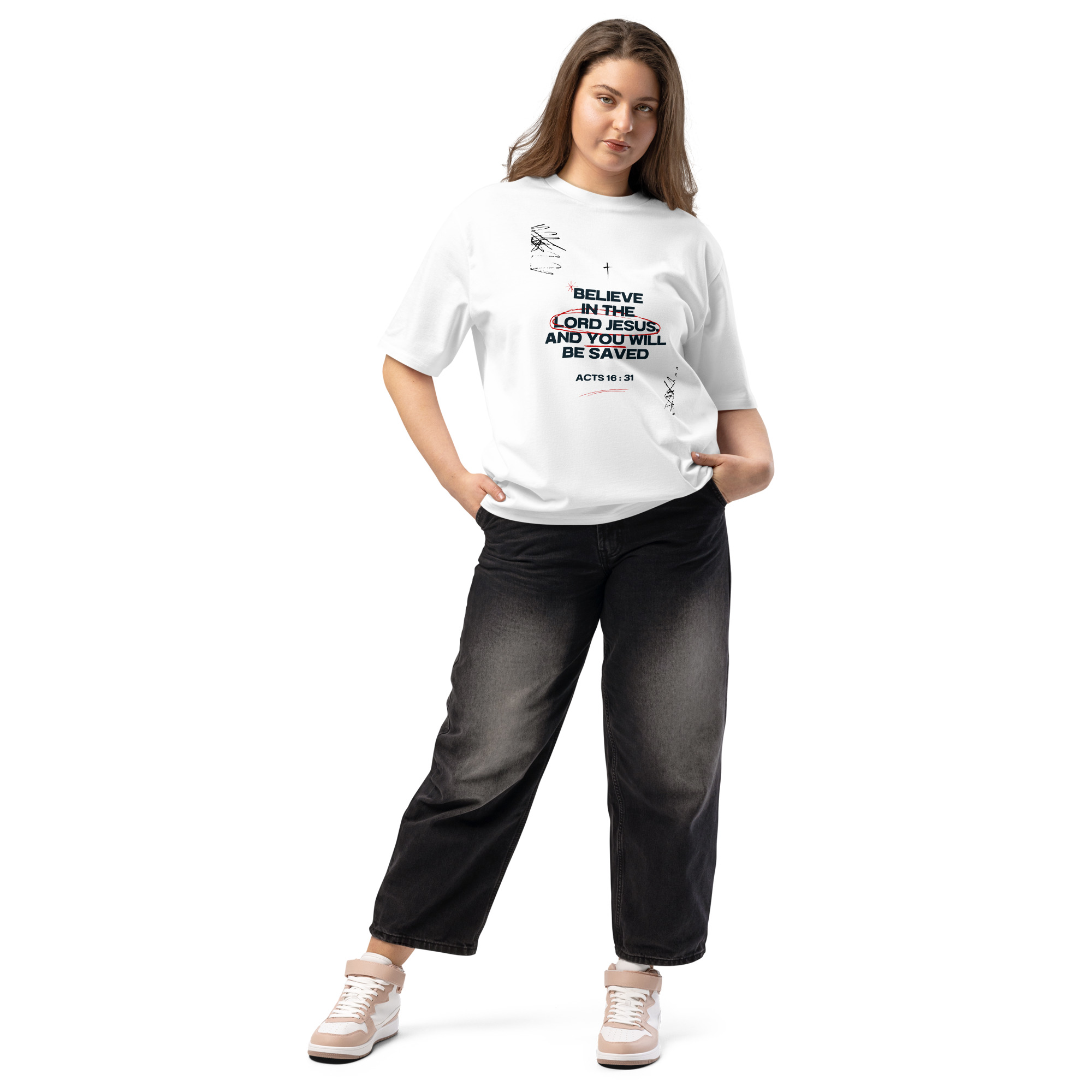 Woman wearing a white T-shirt with a biblical quote from Acts 16:31, black jeans, and sneakers, standing with hands in pockets.