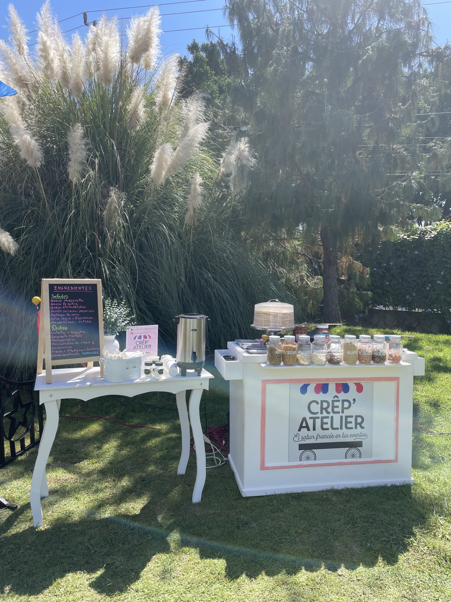 Outdoor crepe station with a colorful menu board, crepe maker, and toppings set up on grass with trees in the background.