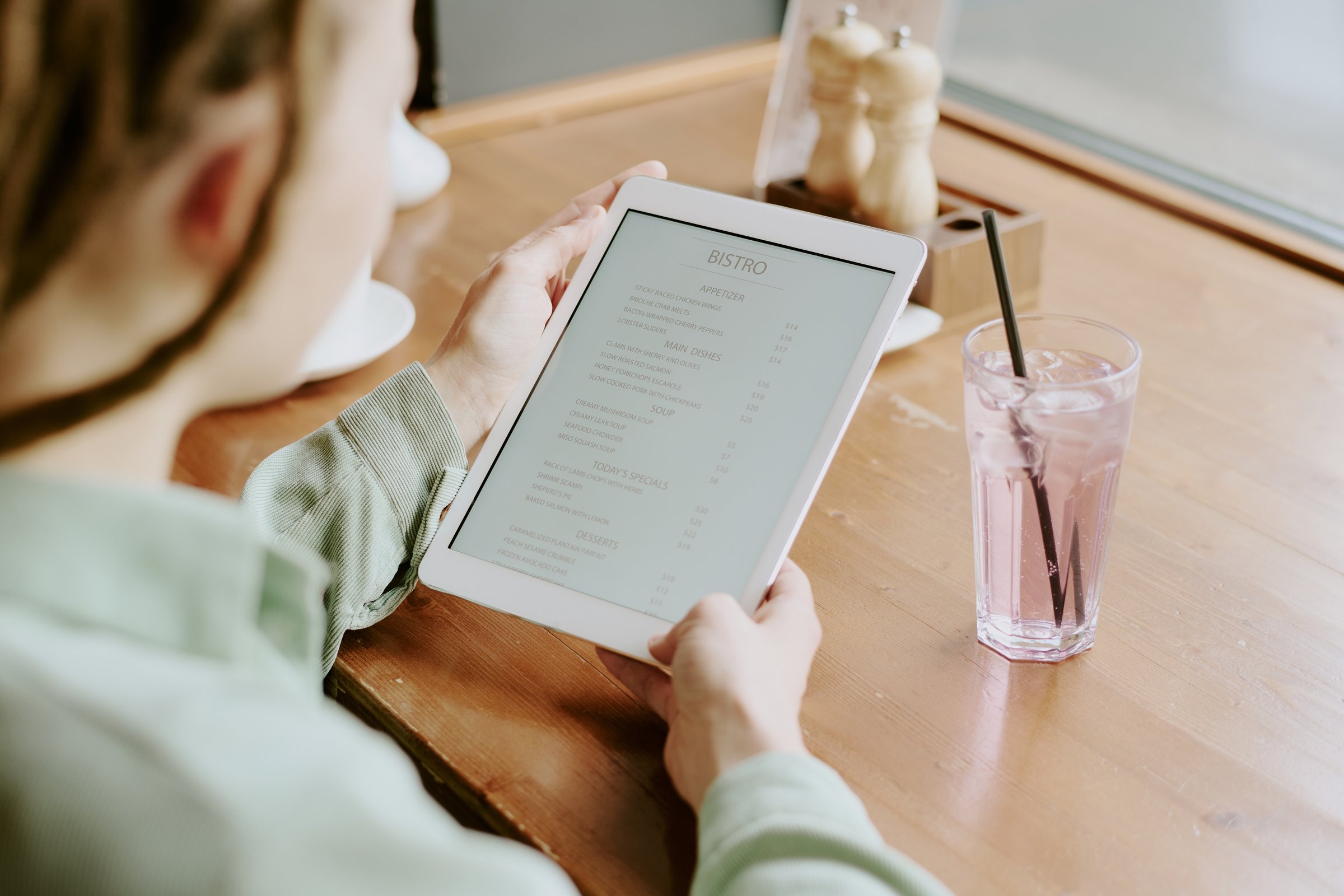 Person holding tablet reviewing digital menu in cozy bistro while drinking refreshment. Hands gripping device revealing tranquil dining setting with glass of beverage