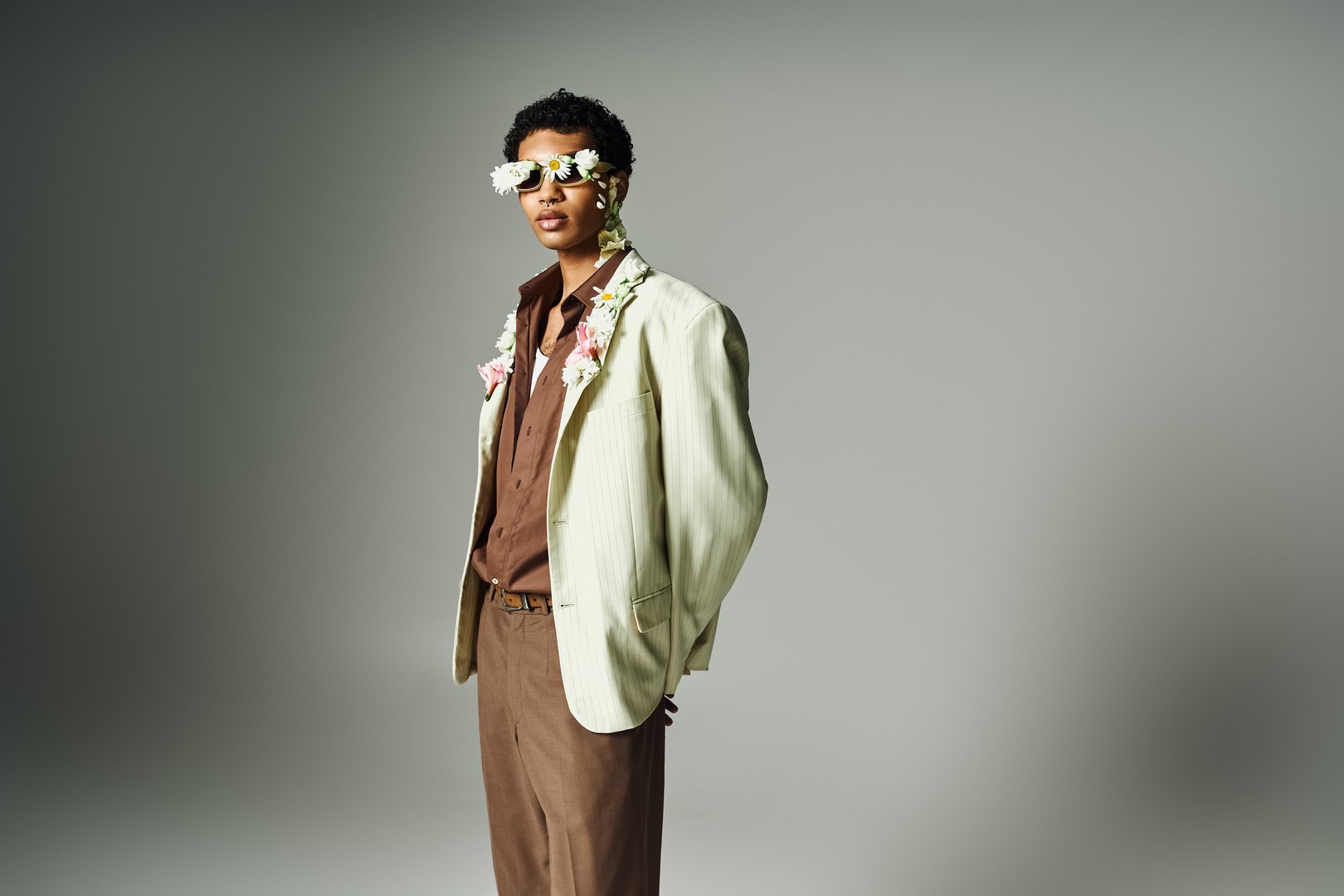 A young African American man poses in a stylish blazer, adorned with flowers around his neck and face, against a grey background.