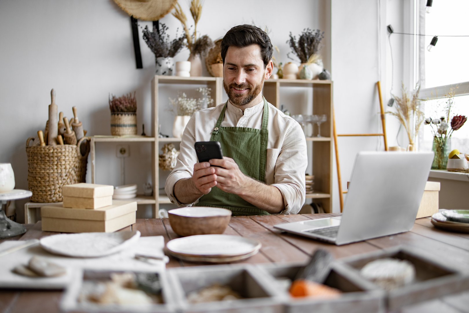 Successful guy store employee with ceramic tableware shoots photos on smartphone. Charismatic Caucasian man seller in souvenir shop takes pictures of goods at phone for posting on social networks.